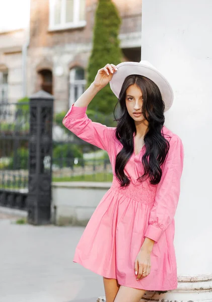 Street style, fashion conception: elegant woman wearing trendy pink dress, white color hat, posing in street of city.