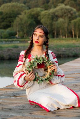 Genç bir esmer Slav kadın bir gölün arka planında elinde çelenkle oturuyor.