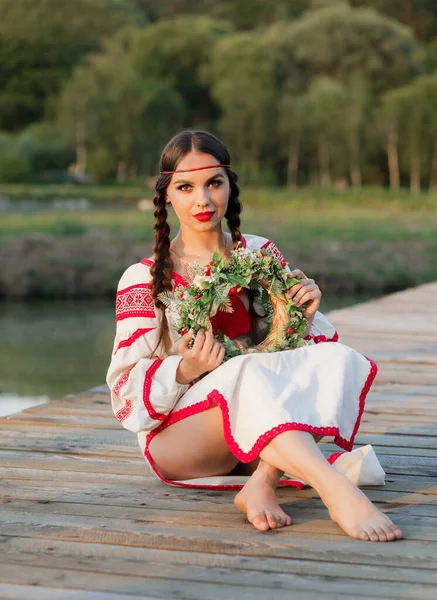 Genç bir esmer Slav kadın bir gölün arka planında elinde çelenkle oturuyor.