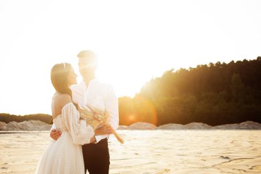 Luxury bride in a white dress ivory shade. Sweet couple is embracing tenderly against the background of white sand at sunset