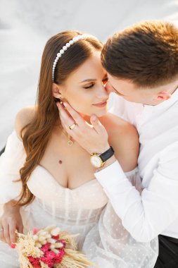 Gentle portrait of the newlyweds on the background of white sand. A man kisses his wife on the cheek. Soft light, natural colors