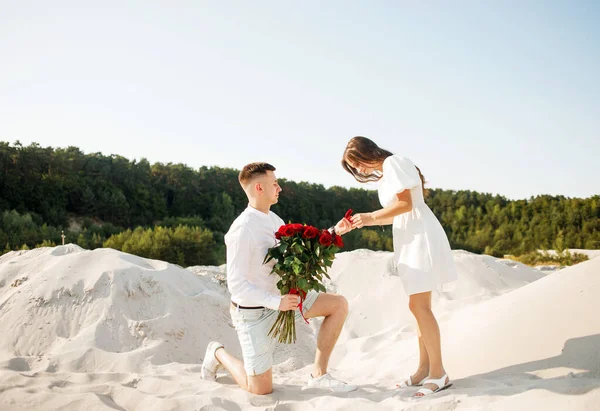 Photo of man making proposal to his amazed woman with ring in gift box while walking on sunny beach