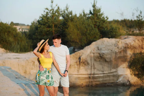 A young couple is relaxing on the shore of a lake, sitting on the sand in summer sunny weather at sunset.