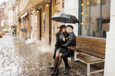 Young couple sitting near a cafe on a bench under an umbrella in rainy weather. Concept of love, romance and passion.