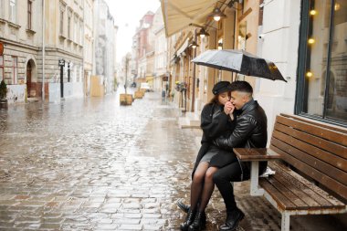 Young couple sitting near a cafe on a bench under an umbrella in rainy weather. Concept of love, romance and passion.