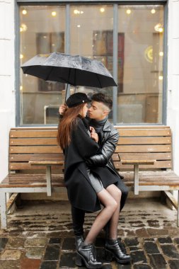 Young couple sitting near a cafe on a bench under an umbrella in rainy weather. Concept of love, romance and passion. 
