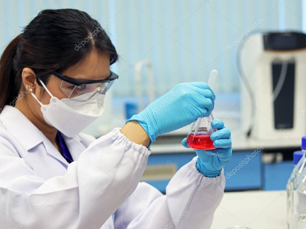 The scientist wears PPE and holds an Erlenmeyer flask laboratory, with red solvent solution from reaction methyl orange indicator acid titration experiment, analysis compounding in wastewater sample.