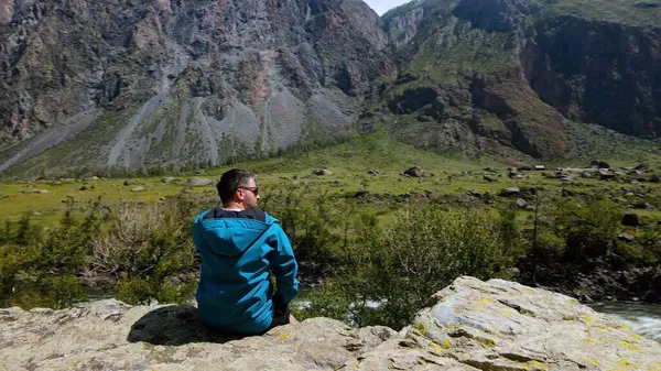 Um homem com óculos senta-se à beira de um penhasco usando um anoraque azul. Katu-Yaryk desfiladeiro Chulyshman Valley. Altai... — Fotografia de Stock