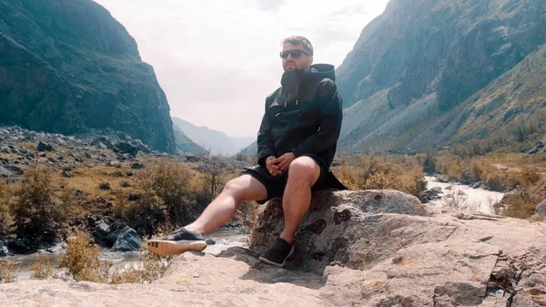 Um homem com óculos senta-se na beira de um penhasco usando um anoraque preto. Katu-Yaryk desfiladeiro Chulyshman Valley. Altai... — Fotografia de Stock