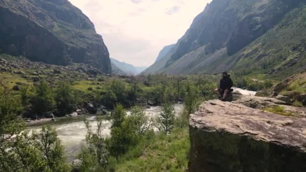 Uçurumun kenarında oturan bir adam siyah bir anorak giyiyor. Katu-Yaryk Kanyonu Chulyshman Vadisi. Altai. — Stok video
