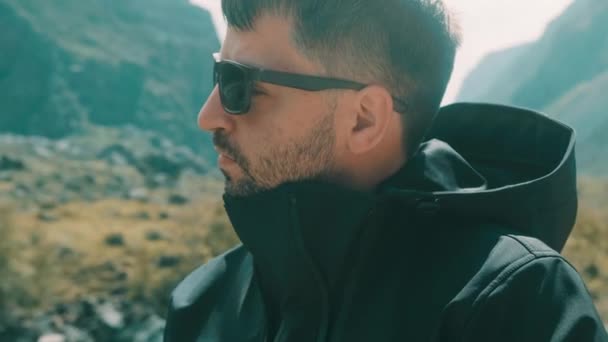 Un homme avec des lunettes est assis sur le bord d'une falaise portant un anorak noir. Katu-Yaryk canyon Chulyshman Valley. Altaï — Video