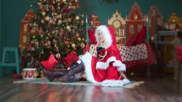 Noel Baba 'nın Noel Baba kıyafetini hediyelerle süsleyen tatlı kız. Yeni yıl elfi. Yere oturur, saçlarını düzeltir. — Stok video