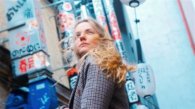 Mavi ceketli bir kız kameraya bakarak caddede yürüyor. Duvarlarda Japonca karakterler