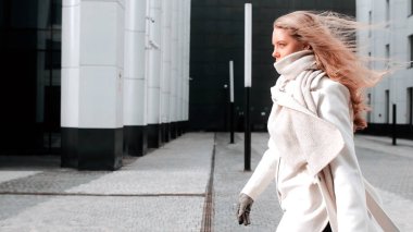 Beyaz önlüklü ve eşarplı bir kız sokakta yürüyor, gülümsüyor. Arka planda iş merkezi var. Başarı