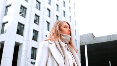 Beyaz önlüklü ve eşarplı bir kız sokakta yürüyor, gülümsüyor. Arka planda iş merkezi var. Başarı