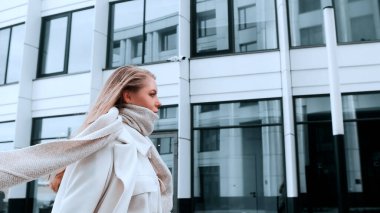 Beyaz önlüklü ve eşarplı bir kız sokakta yürüyor, gülümsüyor. Arka planda iş merkezi var. Başarı