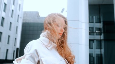 Beyaz önlüklü ve eşarplı bir kız onun kalın saçlarını düzeltir. Güçlü rüzgar. Arka planda iş merkezi var. Başarı