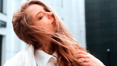 Beyaz önlüklü ve eşarplı bir kız onun kalın saçlarını düzeltir. Güçlü rüzgar. Arka planda iş merkezi var. Başarı