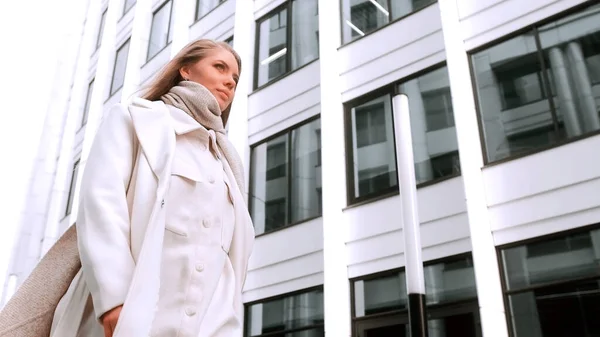 Po ulici kráčí dívka v bílém kabátě a šále a usmívá se. Obchodní centrum v pozadí. Úspěch — Stock fotografie