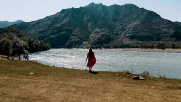Jeune femme en robe rouge se tient près de l'eau incroyable et vue sur la montagne. Drone foootage — Video