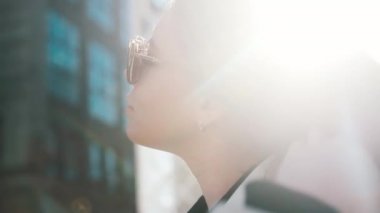 A portrait of a girl with glasses in the bright sun straightens her gorgeous blond hair with her hand, a busy city in the background. Close-up, fashion