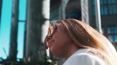 A portrait of a girl with glasses in the bright sun straightens her gorgeous blond hair with her hand, a busy city in the background. Close-up, fashion