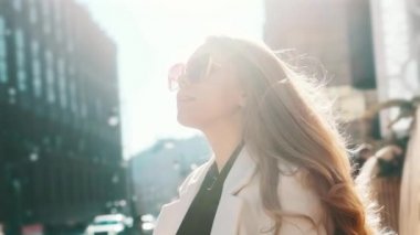 A portrait of a girl with glasses in the bright sun straightens her gorgeous blond hair with her hand, a busy city in the background. Close-up, fashion