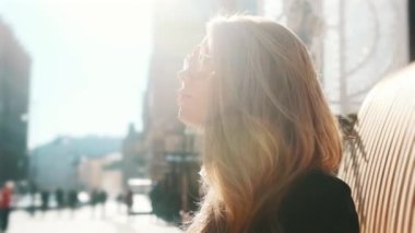 A blonde girl is basking in the bright summer sun on the street, a busy city on the background. Close-up, fashion