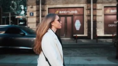 A girl in glasses and a white coat walks through the business district of the city. Against the backdrop of luxury cars and apartments