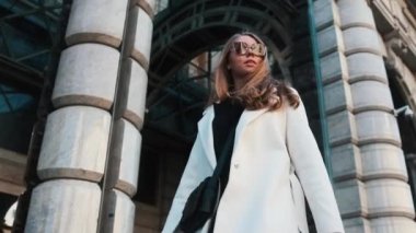 A girl in glasses and a white coat walks through the business district of the city. Against the backdrop of luxury cars and apartments
