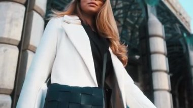 A girl in glasses and a white coat walks through the business district of the city. Against the backdrop of luxury cars and apartments