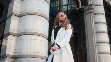 A girl in glasses and a white coat walks through the business district of the city. Against the backdrop of luxury cars and apartments