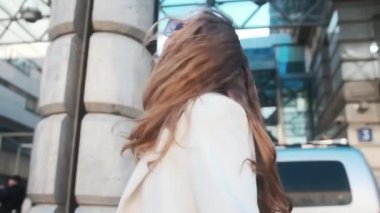 A girl in glasses and a white coat walks through the business district of the city. Against the backdrop of luxury cars and apartments