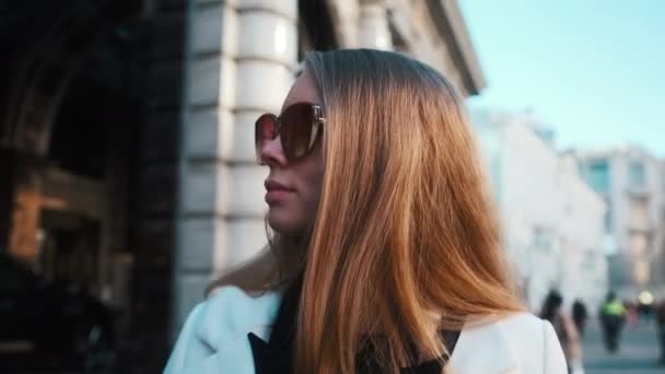 A girl in glasses and a white coat walks through the business district of the city. Against the backdrop of luxury cars and apartments — Vídeo de Stock