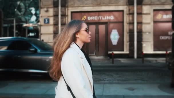 A girl in glasses and a white coat walks through the business district of the city. Against the backdrop of luxury cars and apartments — Vídeo de Stock