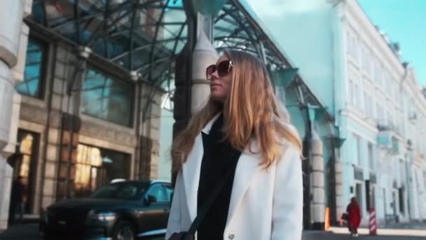 A girl in glasses and a white coat walks through the business district of the city. Against the backdrop of luxury cars and apartments — Vídeo de Stock
