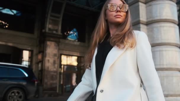 A girl in glasses and a white coat walks through the business district of the city. Against the backdrop of luxury cars and apartments — Stok video