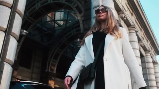 A girl in glasses and a white coat walks through the business district of the city. Against the backdrop of luxury cars and apartments — Stockvideo