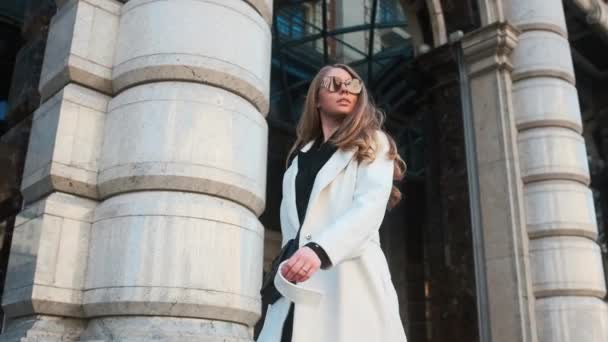 A girl in glasses and a white coat walks through the business district of the city. Against the backdrop of luxury cars and apartments — Stock videók