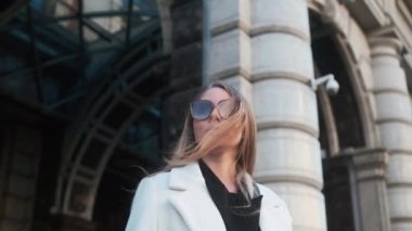 A girl in glasses and a white coat stands against the background of a hotel in the city center and adjusts her glasses
