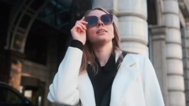A girl in glasses and a white coat stands against the background of a hotel in the city center and adjusts her glasses