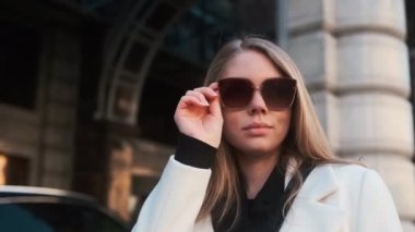 A girl in glasses and a white coat stands against the background of a hotel in the city center and adjusts her glasses