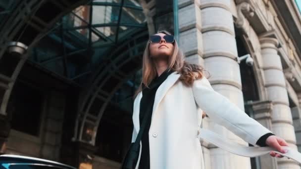 Ένα κορίτσι με γυαλιά και ένα λευκό παλτό στέκεται στο φόντο ενός ξενοδοχείου στο κέντρο της πόλης και κοιτάζει γύρω του. — Αρχείο Βίντεο