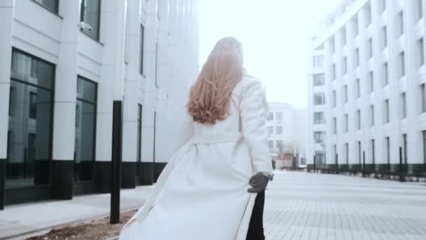 A girl in a white coat and a scarf is walking down the street, smiling. Business center in the background. Success — Stock Video