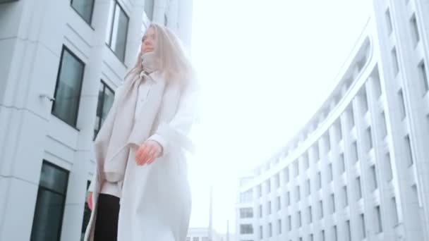 Dziewczyna w białym płaszczu i szaliku idzie ulicą, uśmiechnięta. Centrum biznesowe w tle. Sukces — Wideo stockowe
