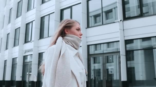 Une fille en manteau blanc et une écharpe marchent dans la rue, souriant. Centre d'affaires en arrière-plan. Succès — Video