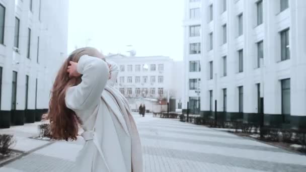 A girl in a white coat and a scarf straightens her thick hair. Strong wind. Business center in the background. Success — Stockvideo