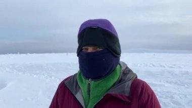 A warmly dressed man in a mask that covers his entire face except for his eyes looks into the camera. Close-up on the eyes. Winter hike. Harsh northern nature. Beautiful winter landscape.