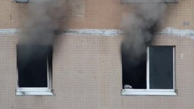 Gray thick smoke billows from two windows of an apartment building. Extinguishing a fire in a high-rise building. Fire in the apartment. A firefighter shines a lantern in a dark window.