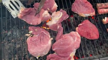 A piece of raw red meat is turned over to the other side for better frying with iron tongs and a spatula. Cooking barbecue and steaks on a charcoal grill. Summer holidays and vacations. Picnic in the country.